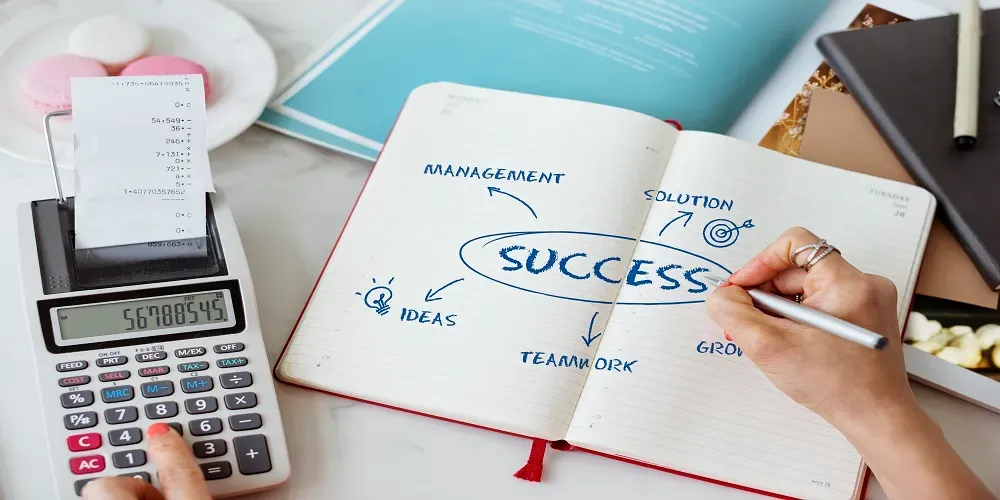 A person writing the word "success" in a notebook with arrows pointing to words like management, ideas, teamwork, growth, and solution, while using a calculator nearby.